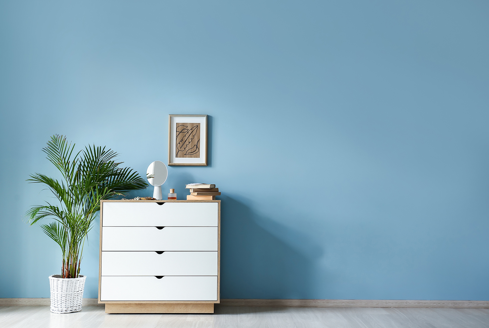 Zimmer mit hellblauer Wandfarbe, weißer Kommode links und einer Palme daneben.