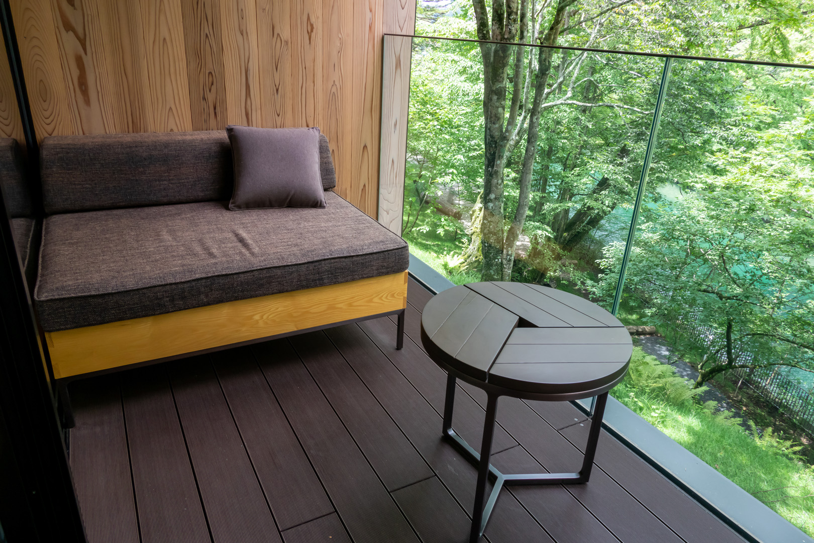 Balkon mit dunklem Holzboden, Glaswand, Sessel, kleinem runden Holztisch und Blick in einen Wald