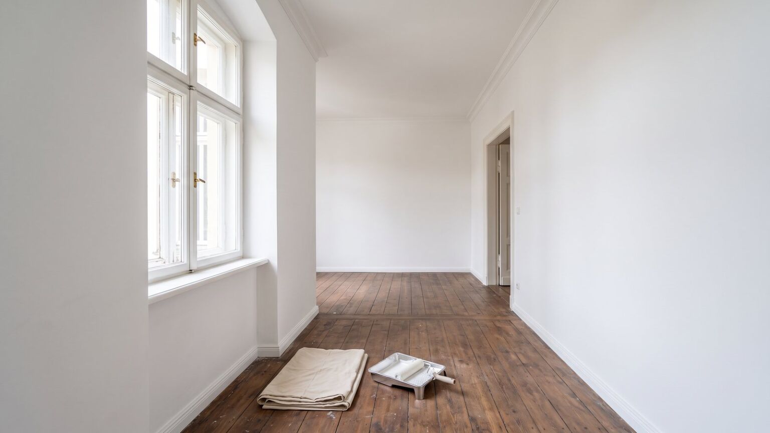 Frisch weiß gestrichenes Altbauzimmer mit tiefer Fensterlaibung, auf dem Dielenboden ein gefaltetes Abdeckvlies und eine Farbrolle in der Wanne